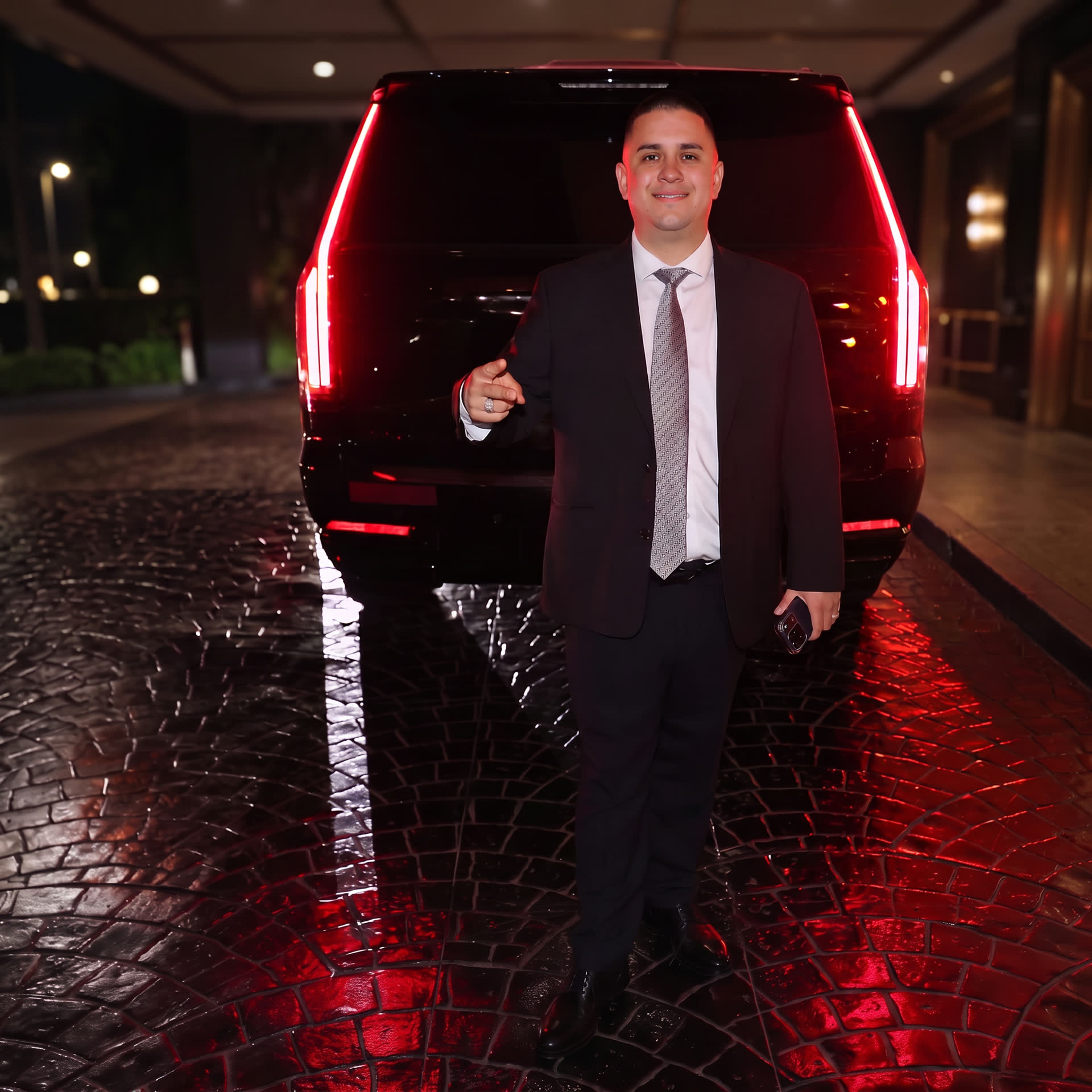 Professional chauffeur in a suit welcoming guests beside a black Cadillac Escalade at a hotel entrance at night